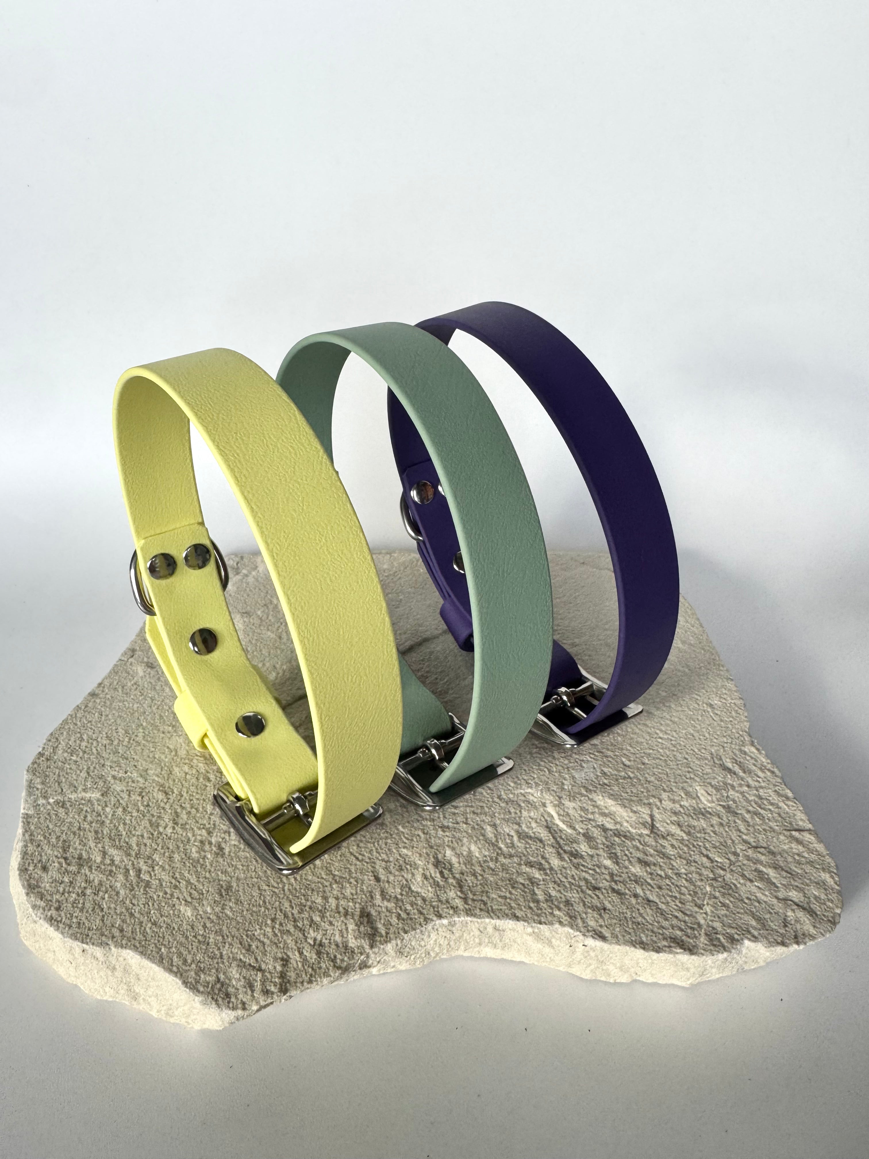 Three dog collars in yellow, green, and purple on a stone surface with a white background